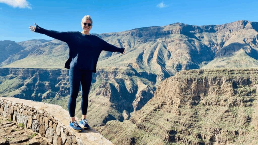A smiling person standing on a stone ledge with arms outstretched, overlooking a vast, sunlit mountain canyon under a clear blue sky.